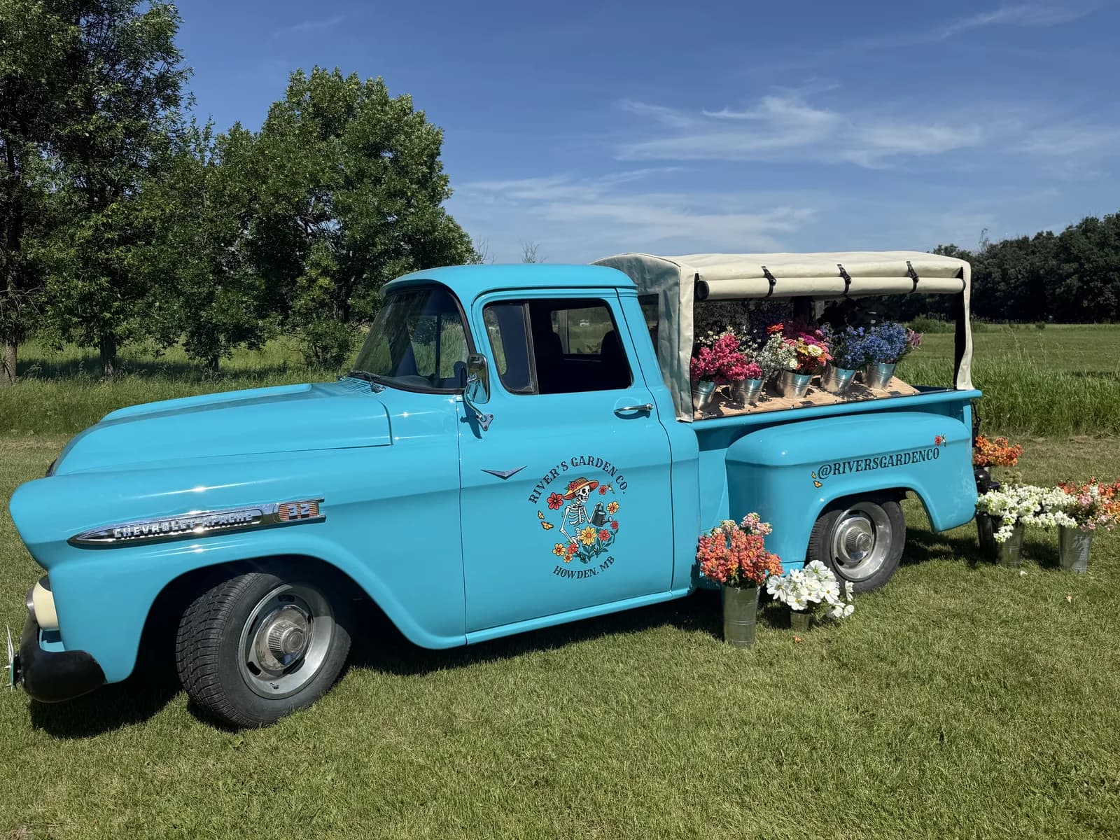 The full Flower Truck — Tartan Turquoise 1958 Chevy Apache with white canopy and the River's Garden Co. skeleton logo on the door — parked in a field with the day's stems loaded in back.