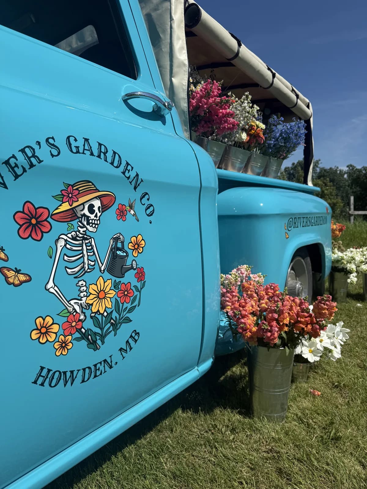 Close-up of the hood — the River's Garden Co. skeleton logo arced beneath 'HOWDEN, MB', teal paint catching the light.